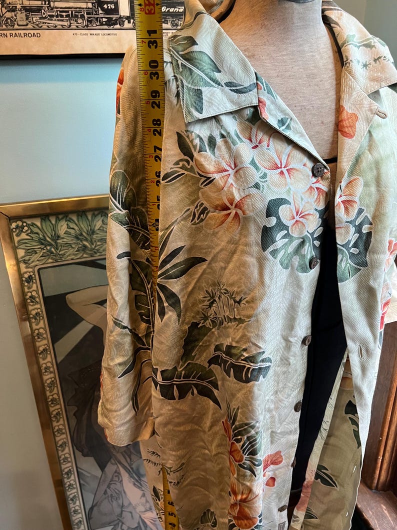 May include: A light-colored button-down shirt with a tropical floral pattern featuring hibiscus flowers and large green leaves. The shirt is measured at approximately 30 inches in length. The shirt is on a mannequin.