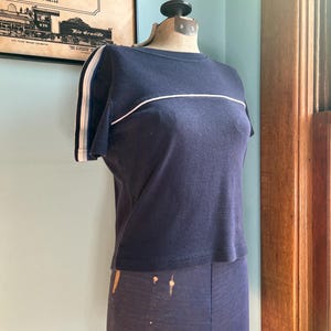 K&ouml;nnte beinhalten: Marineblaues T-Shirt mit wei&szlig;en Streifen auf den &Auml;rmeln und &uuml;ber der Brust. Das Shirt wird auf einer Schaufensterpuppe pr&auml;sentiert. Im Hintergrund befindet sich ein Vintage-Poster.