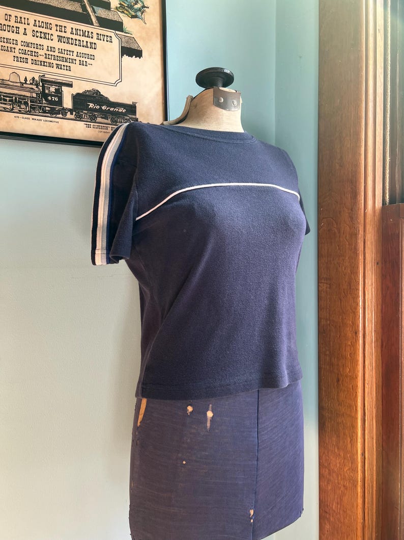 K&ouml;nnte beinhalten: Marineblaues T-Shirt mit wei&szlig;en Streifen auf den &Auml;rmeln und &uuml;ber der Brust. Das Shirt wird auf einer Schaufensterpuppe pr&auml;sentiert. Im Hintergrund befindet sich ein Vintage-Poster.