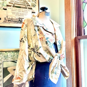 May include: A beige floral print shirt with a tie front is displayed on a mannequin. The shirt features a pattern of orange and pink flowers with green leaves. The mannequin is wearing a dark blue garment underneath. The background includes vintage artwork and a window.