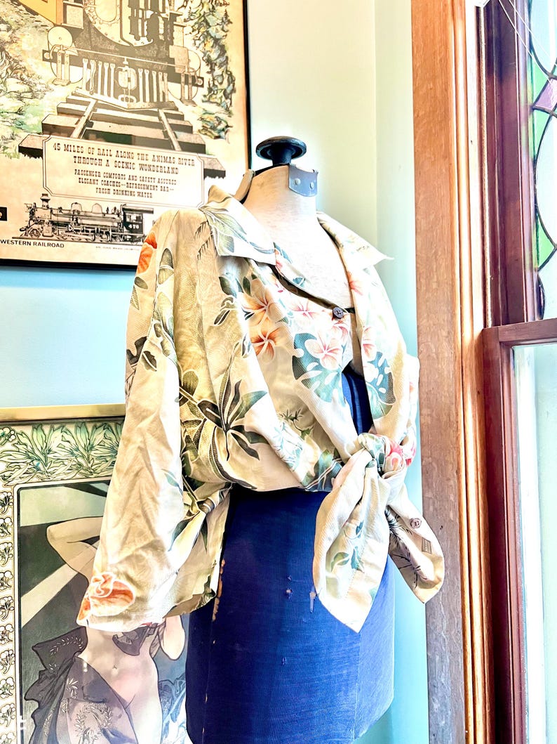 May include: A beige floral print shirt with a tie front is displayed on a mannequin. The shirt features a pattern of orange and pink flowers with green leaves. The mannequin is wearing a dark blue garment underneath. The background includes vintage artwork and a window.