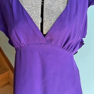 May include: A vibrant purple dress with a deep V-neckline and short, ruffled sleeves. The dress features a gathered waist and a flowing, lightweight fabric, perfect for warm weather. The dress is displayed on a mannequin.