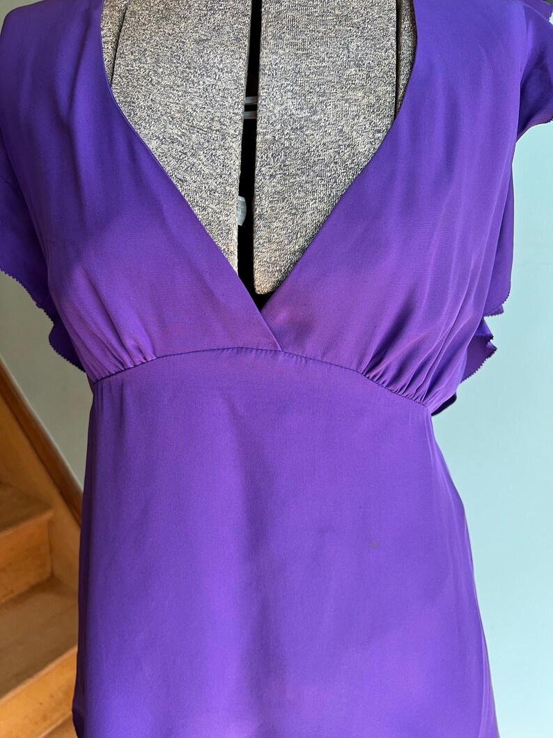 May include: A vibrant purple dress with a deep V-neckline and short, ruffled sleeves. The dress features a gathered waist and a flowing, lightweight fabric, perfect for warm weather. The dress is displayed on a mannequin.