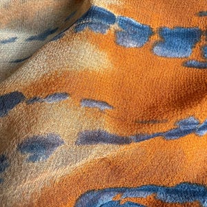 Peut inclure: Gros plan sur un tissu au design abstrait. Le tissu pr&eacute;sente un m&eacute;lange de teintes orange, bleu et beige. Le motif comprend des formes bleues irr&eacute;guli&egrave;res sur un fond orange, avec des accents beiges. Le tissu semble doux et l&eacute;ger.
