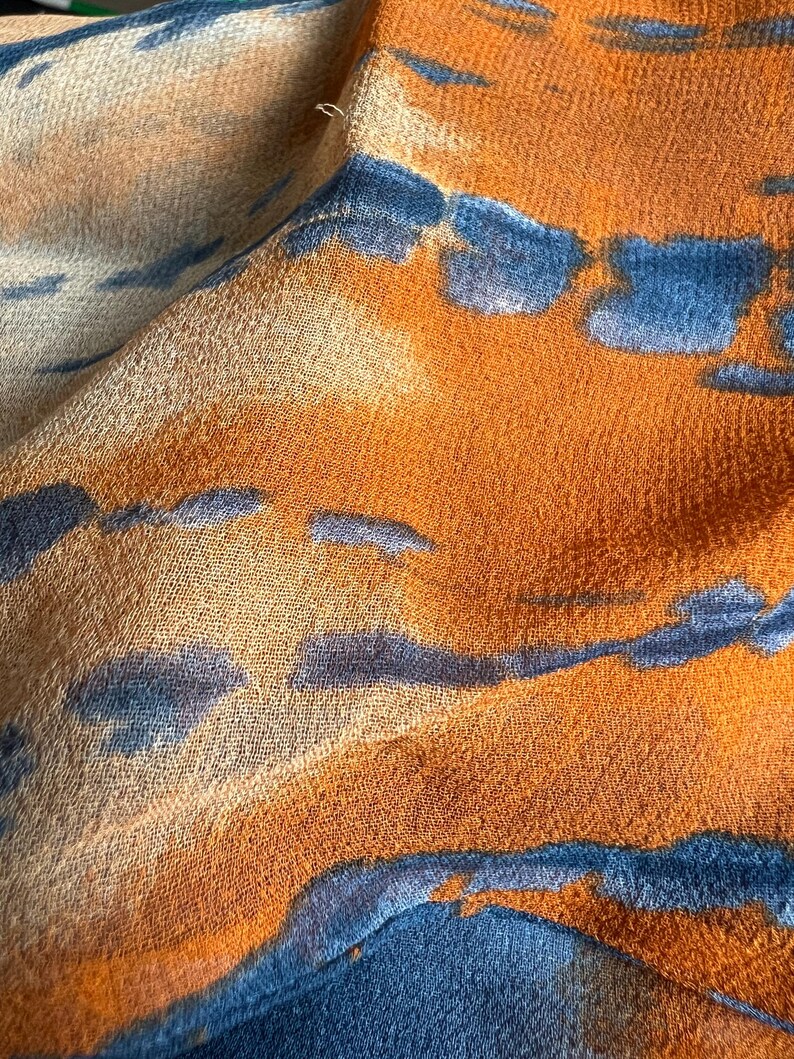 Peut inclure: Gros plan sur un tissu au design abstrait. Le tissu pr&eacute;sente un m&eacute;lange de teintes orange, bleu et beige. Le motif comprend des formes bleues irr&eacute;guli&egrave;res sur un fond orange, avec des accents beiges. Le tissu semble doux et l&eacute;ger.