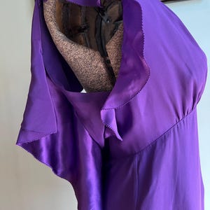 May include: A vibrant purple dress with a flowing, ruffled shoulder detail. The dress is made of a shiny, satin-like material. The dress is displayed on a mannequin.