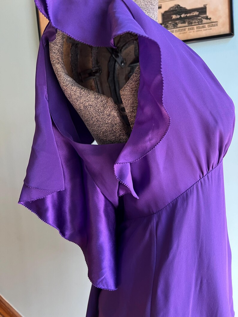 May include: A vibrant purple dress with a flowing, ruffled shoulder detail. The dress is made of a shiny, satin-like material. The dress is displayed on a mannequin.