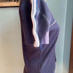 K&ouml;nnte beinhalten: Ein marineblaues Kurzarmshirt mit wei&szlig;en und hellblauen Streifen an den &Auml;rmeln. Das Kleidungsst&uuml;ck wird auf einer Schaufensterpuppe pr&auml;sentiert. Das Oberteil hat einen Rundhalsausschnitt und eine taillierte Form. Der Stoff scheint ein weiches Strickmaterial zu sein.