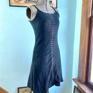 May include: A black sleeveless dress featuring a vertical row of silver-toned metal hook-and-eye closures running down the center front. The garment has thin spaghetti straps, a scoop neckline, and a fitted bodice that flares slightly at the hem.