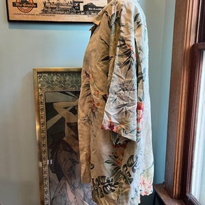 May include: A beige button-down shirt with a tropical floral pattern in shades of green, red, and orange. The shirt is displayed on a mannequin. The shirt has long sleeves and a collared neckline. The background includes a vintage poster and artwork.