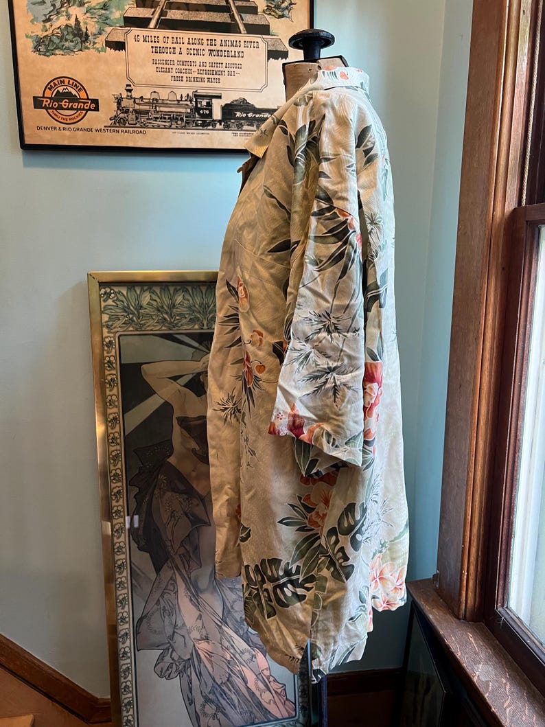 May include: A beige button-down shirt with a tropical floral pattern in shades of green, red, and orange. The shirt is displayed on a mannequin. The shirt has long sleeves and a collared neckline. The background includes a vintage poster and artwork.