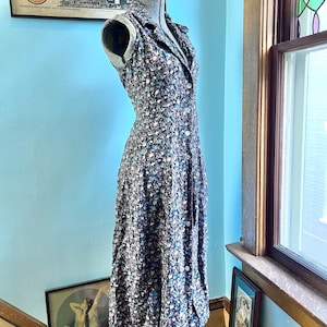 May include: A sleeveless midi dress featuring a dark base with a dense floral pattern in shades of blue, purple, and white. The dress has a collared neckline, a button-down front with white buttons, and a fitted bodice that flows into a skirt.