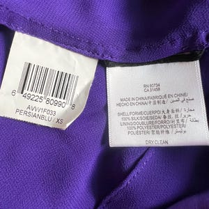 May include: Close-up of a purple garment with attached tags. One tag displays a barcode and the text "PERSIANBLU/XS". Another tag includes the text "MADE IN CHINA" and fabric composition details. The garment is made of silk and polyester.