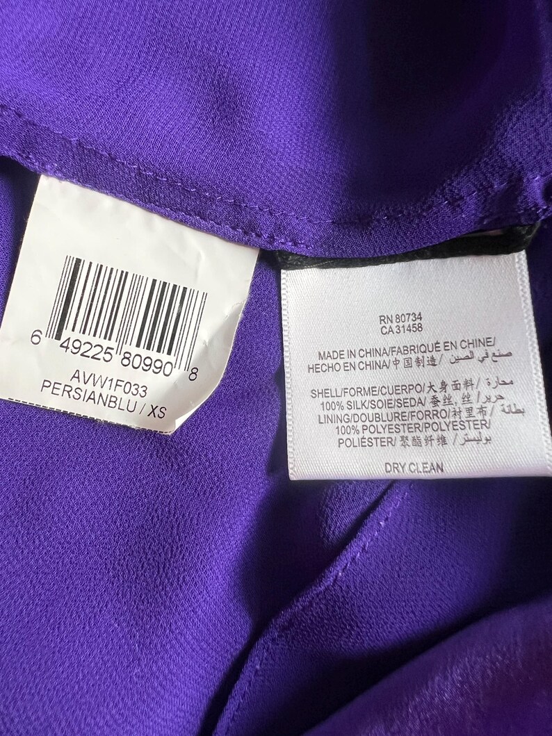 May include: Close-up of a purple garment with attached tags. One tag displays a barcode and the text "PERSIANBLU/XS". Another tag includes the text "MADE IN CHINA" and fabric composition details. The garment is made of silk and polyester.