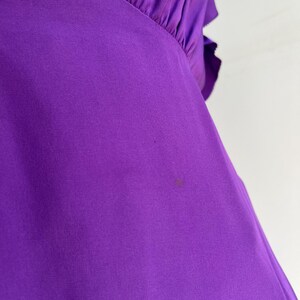 May include: Close-up of a vibrant purple garment, possibly a blouse or dress, with a gathered neckline and ruffled sleeve detail. The fabric appears to be a smooth, flowing material. A small stain is visible on the fabric.