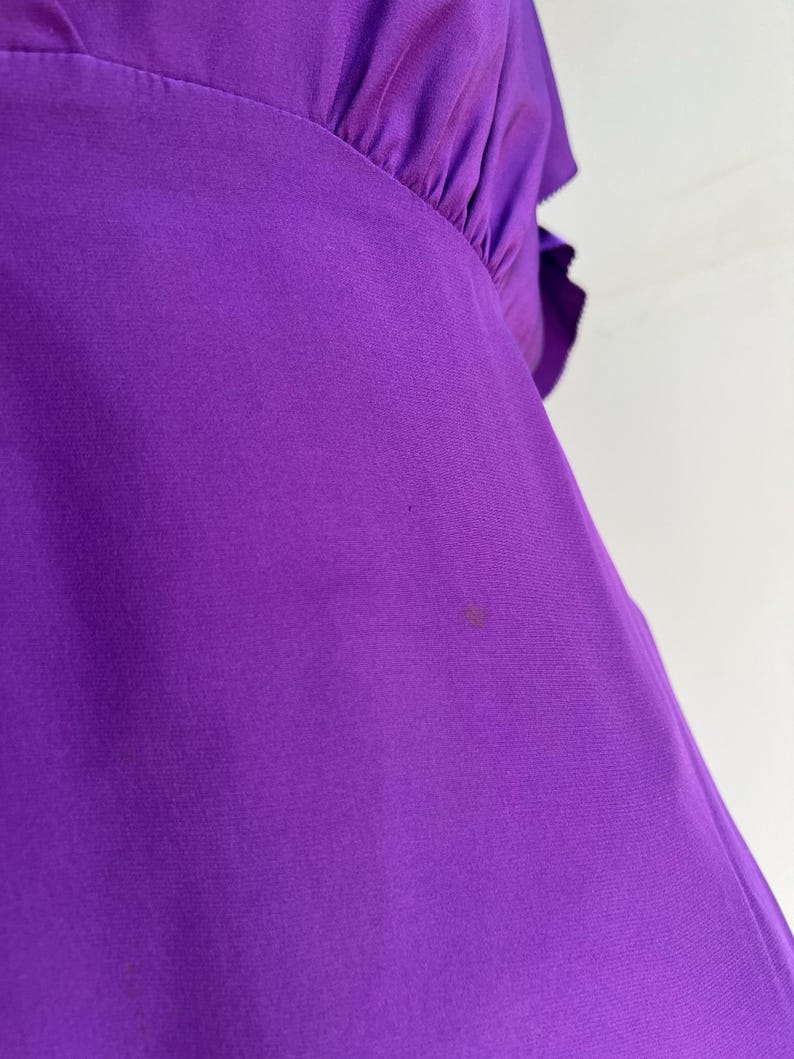 May include: Close-up of a vibrant purple garment, possibly a blouse or dress, with a gathered neckline and ruffled sleeve detail. The fabric appears to be a smooth, flowing material. A small stain is visible on the fabric.