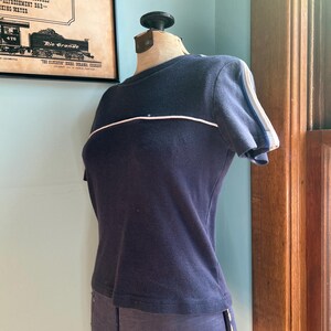 K&ouml;nnte beinhalten: Ein marineblaues Kurzarm-Shirt mit einem wei&szlig;en Streifen &uuml;ber der Brust und wei&szlig;en und blauen Streifen auf den &Auml;rmeln. Das Shirt wird auf einer Schaufensterpuppe pr&auml;sentiert. Teil einer Bekleidungskollektion.