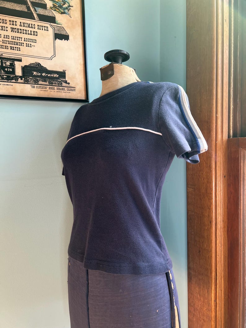 K&ouml;nnte beinhalten: Ein marineblaues Kurzarm-Shirt mit einem wei&szlig;en Streifen &uuml;ber der Brust und wei&szlig;en und blauen Streifen auf den &Auml;rmeln. Das Shirt wird auf einer Schaufensterpuppe pr&auml;sentiert. Teil einer Bekleidungskollektion.