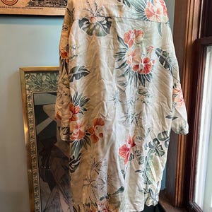 May include: A beige button-down shirt with a tropical floral pattern featuring orange and red hibiscus flowers and green leaves. The shirt is hanging on a display, showcasing the back of the garment. The shirt has a classic collar and long sleeves.