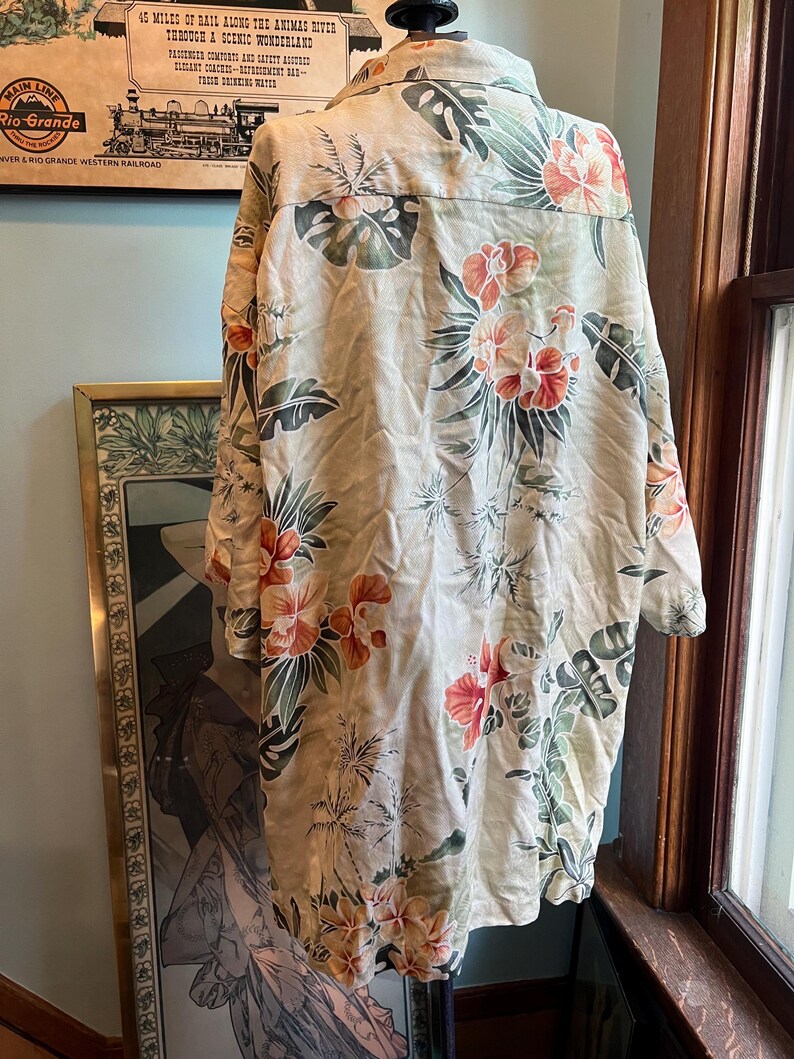 May include: A beige button-down shirt with a tropical floral pattern featuring orange and red hibiscus flowers and green leaves. The shirt is hanging on a display, showcasing the back of the garment. The shirt has a classic collar and long sleeves.