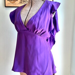 May include: A vibrant purple blouse with a deep V-neck and ruffled cap sleeves. The top is made of a smooth, flowing fabric and has a slightly flared silhouette. The blouse is displayed on a mannequin.