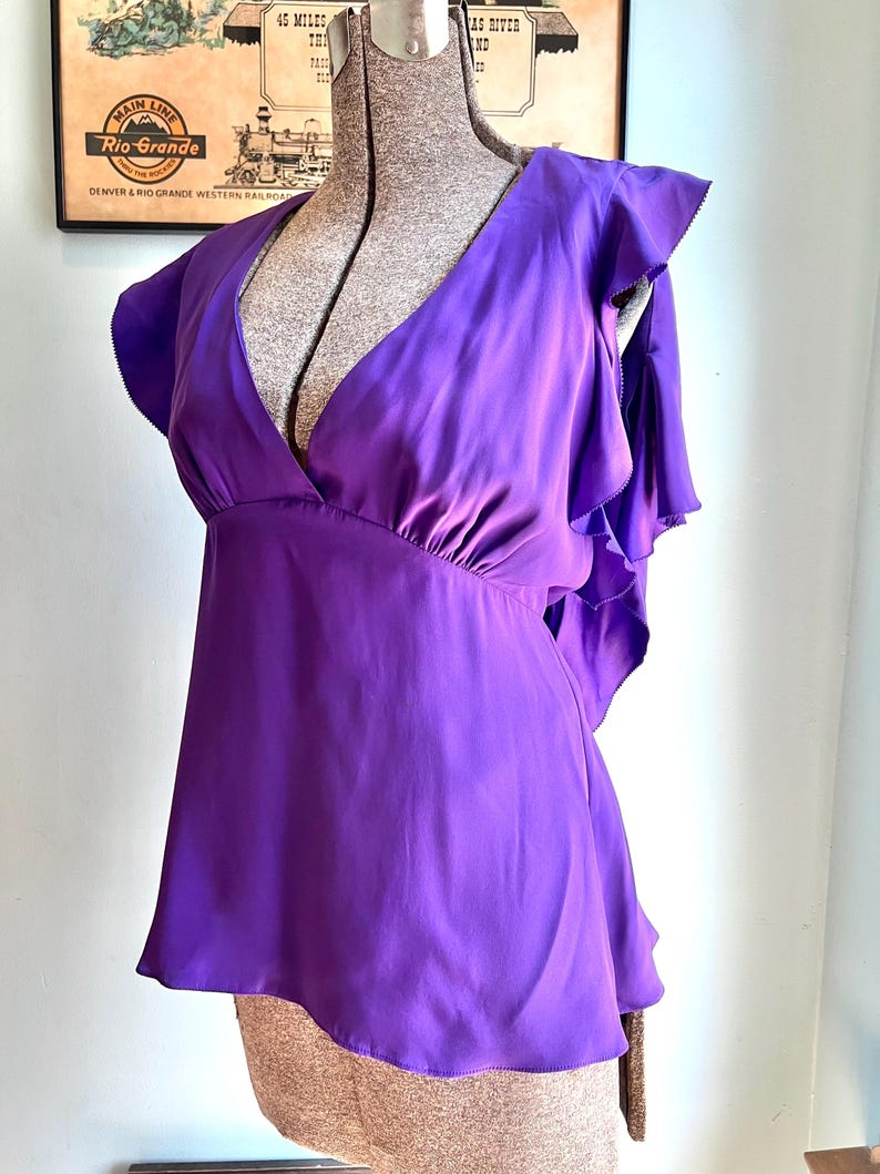 May include: A vibrant purple blouse with a deep V-neck and ruffled cap sleeves. The top is made of a smooth, flowing fabric and has a slightly flared silhouette. The blouse is displayed on a mannequin.