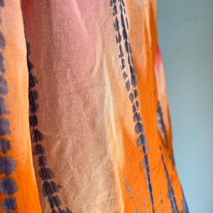 Peut inclure: Gros plan sur un tissu fluide avec un motif tie-dye. Les couleurs comprennent l'orange, le rose et le bleu, avec des lignes verticales bleu fonc&eacute;. Le tissu semble l&eacute;ger et semi-transparent, sugg&eacute;rant une &eacute;charpe ou un ch&acirc;le.