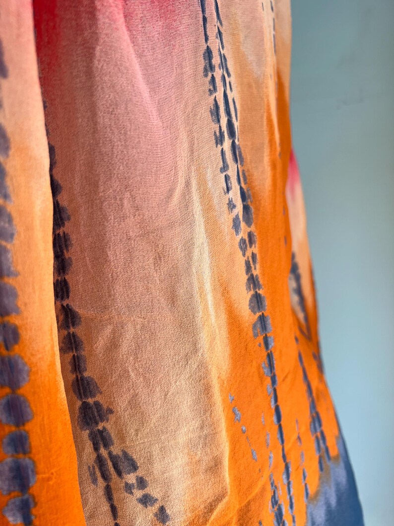 Peut inclure: Gros plan sur un tissu fluide avec un motif tie-dye. Les couleurs comprennent l'orange, le rose et le bleu, avec des lignes verticales bleu fonc&eacute;. Le tissu semble l&eacute;ger et semi-transparent, sugg&eacute;rant une &eacute;charpe ou un ch&acirc;le.