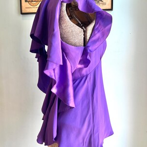 May include: A vibrant purple top with layered ruffles and a draped design. The garment is displayed on a mannequin, showcasing its flowing silhouette and unique texture. The top's color is a rich, saturated purple.