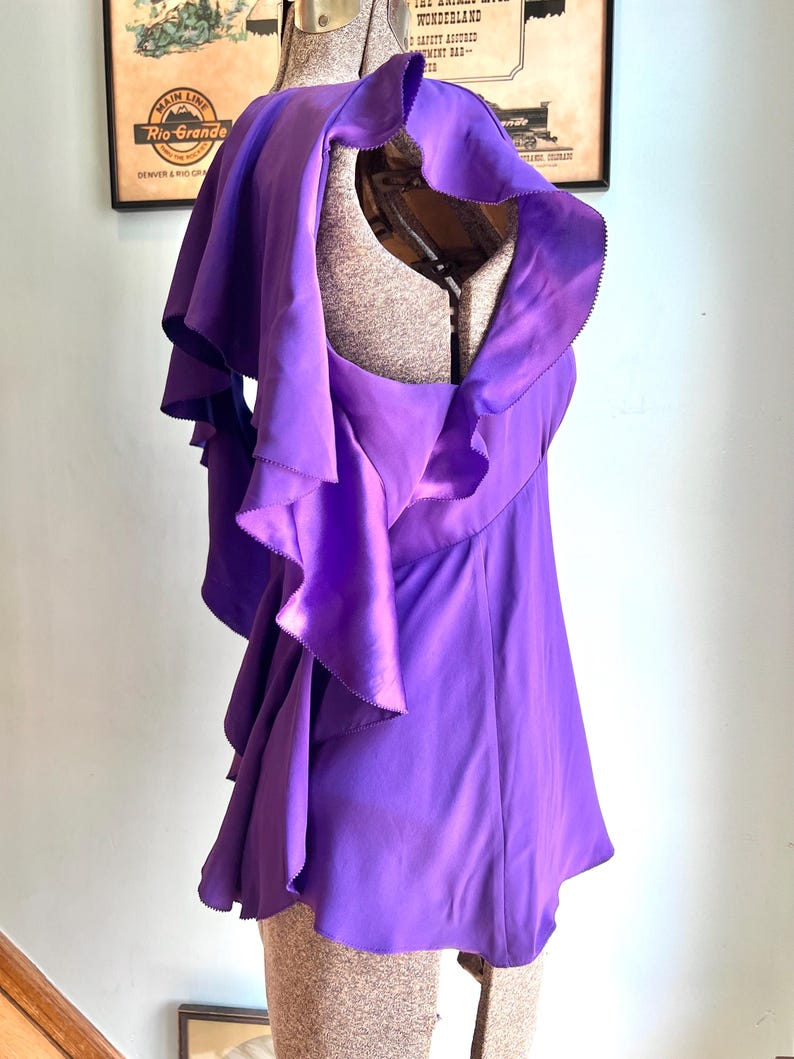May include: A vibrant purple top with layered ruffles and a draped design. The garment is displayed on a mannequin, showcasing its flowing silhouette and unique texture. The top's color is a rich, saturated purple.