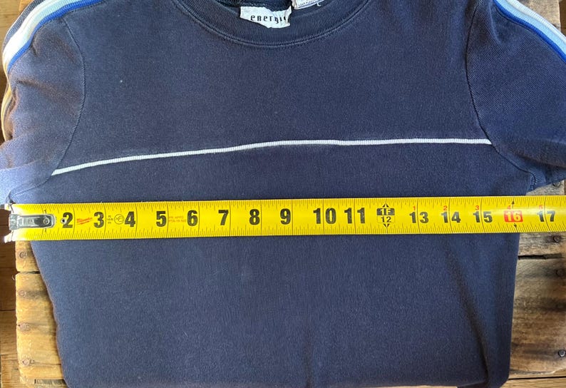 K&ouml;nnte beinhalten: Marineblaues Sweatshirt mit Rundhalsausschnitt und einem wei&szlig;en horizontalen Streifen &uuml;ber der Brust. Die &Auml;rmel haben blaue Streifen. Das Ma&szlig;band zeigt, dass das Hemd etwa 40 cm breit ist. Das Etikett lautet "energy".