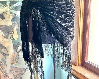 May include: A black shawl with a patterned design and long fringe. The shawl is draped over a mannequin, showcasing its texture and drape. The design appears to be a velvet-like material.