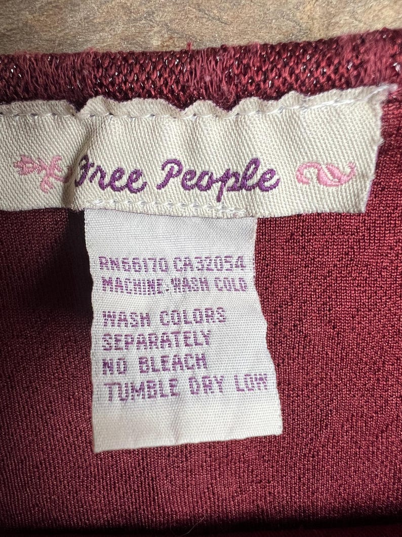 May include: A white care label with the text "free People" in purple lettering, followed by the text "RN66170 CA32054 MACHINE WASH COLD WASH COLORS SEPARATELY NO BLEACH TUMBLE DRY LOW".