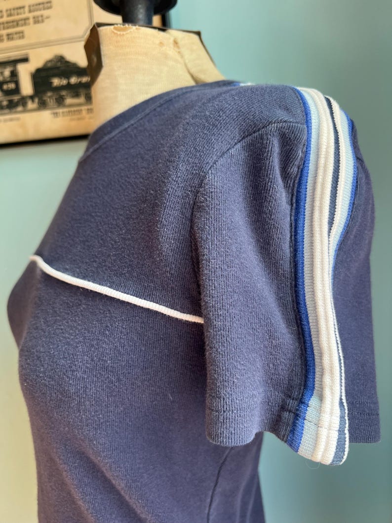 K&ouml;nnte beinhalten: Marineblaues Kurzarm-T-Shirt mit wei&szlig;er Paspelierung und gestreiften Akzenten am &Auml;rmel. Die Streifen umfassen Blau- und Wei&szlig;t&ouml;ne. Das Shirt wird auf einer Schaufensterpuppe pr&auml;sentiert.