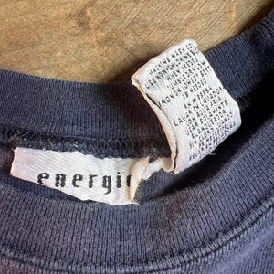 K&ouml;nnte beinhalten: Nahaufnahme eines marineblauen Rundhals-T-Shirts. Ein wei&szlig;es Etikett mit dem Wort "energi" ist in den Kragen eingen&auml;ht. Ein zweites Etikett mit Waschanweisungen ist ebenfalls sichtbar. Das Shirt scheint aus einem weichen, bequemen Material zu sein.