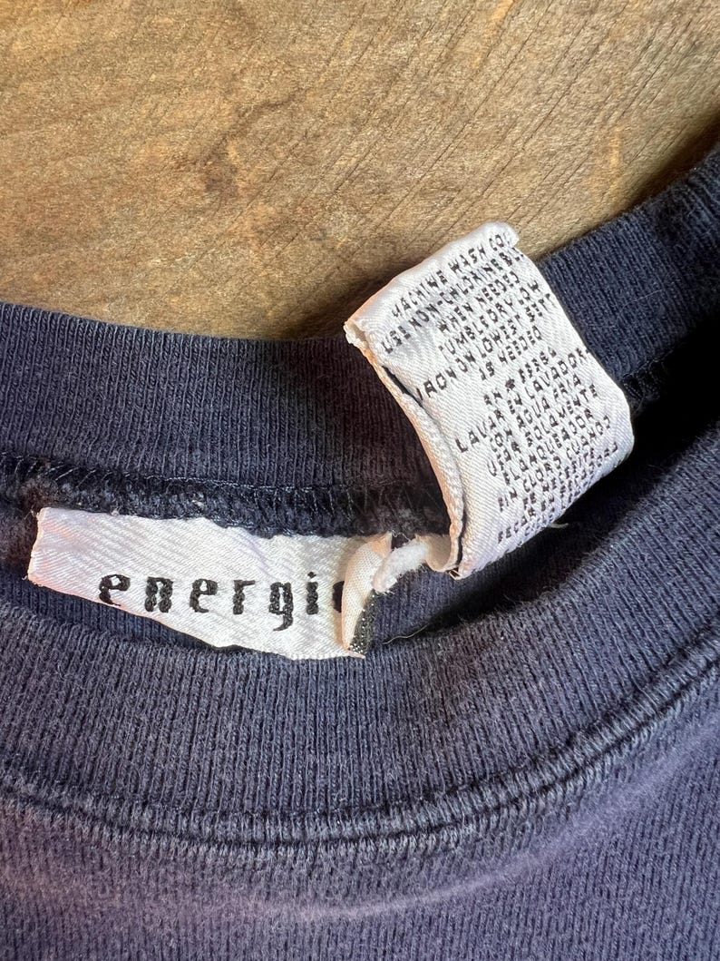 K&ouml;nnte beinhalten: Nahaufnahme eines marineblauen Rundhals-T-Shirts. Ein wei&szlig;es Etikett mit dem Wort "energi" ist in den Kragen eingen&auml;ht. Ein zweites Etikett mit Waschanweisungen ist ebenfalls sichtbar. Das Shirt scheint aus einem weichen, bequemen Material zu sein.