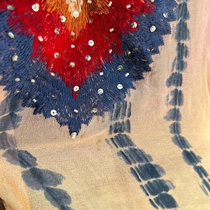 Peut inclure: Gros plan sur un tissu transparent de couleur claire avec un motif brod&eacute; d&eacute;taill&eacute;. La broderie pr&eacute;sente un motif en forme de c&oelig;ur en rouge, orange et bleu, orn&eacute; de paillettes. Des rayures verticales bleu tie-dye descendent le tissu.