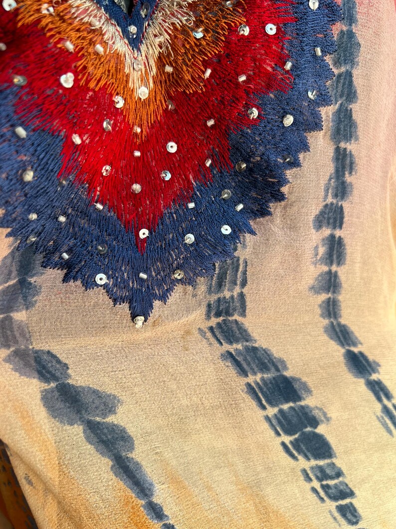 Peut inclure: Gros plan sur un tissu transparent de couleur claire avec un motif brod&eacute; d&eacute;taill&eacute;. La broderie pr&eacute;sente un motif en forme de c&oelig;ur en rouge, orange et bleu, orn&eacute; de paillettes. Des rayures verticales bleu tie-dye descendent le tissu.