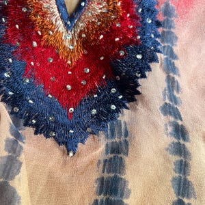 Peut inclure: Gros plan sur un v&ecirc;tement color&eacute; avec un d&eacute;collet&eacute; en V. L'encolure pr&eacute;sente des broderies complexes en rouge, orange et bleu, orn&eacute;es de petites perles scintillantes. Le tissu pr&eacute;sente un motif tie-dye dans les tons p&ecirc;che et bleu.