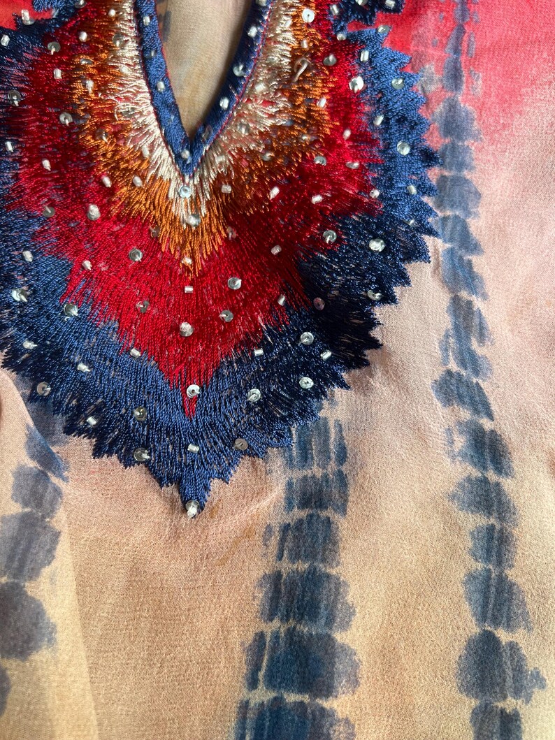 Peut inclure: Gros plan sur un v&ecirc;tement color&eacute; avec un d&eacute;collet&eacute; en V. L'encolure pr&eacute;sente des broderies complexes en rouge, orange et bleu, orn&eacute;es de petites perles scintillantes. Le tissu pr&eacute;sente un motif tie-dye dans les tons p&ecirc;che et bleu.