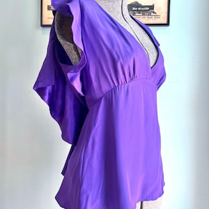 May include: A vibrant purple blouse with a deep V-neck and ruffled cap sleeves. The silky fabric drapes elegantly, and the blouse is displayed on a mannequin. The design features a fitted waist and a flared hem, creating a flattering silhouette.