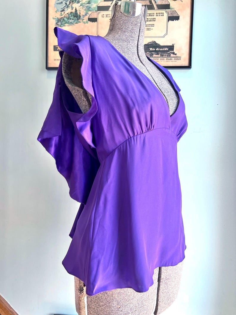 May include: A vibrant purple blouse with a deep V-neck and ruffled cap sleeves. The silky fabric drapes elegantly, and the blouse is displayed on a mannequin. The design features a fitted waist and a flared hem, creating a flattering silhouette.