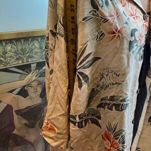 May include: A beige button-down shirt with a tropical floral print in orange, green, and white. The shirt is measured with a yellow tape measure, showing a length of approximately 25 inches.