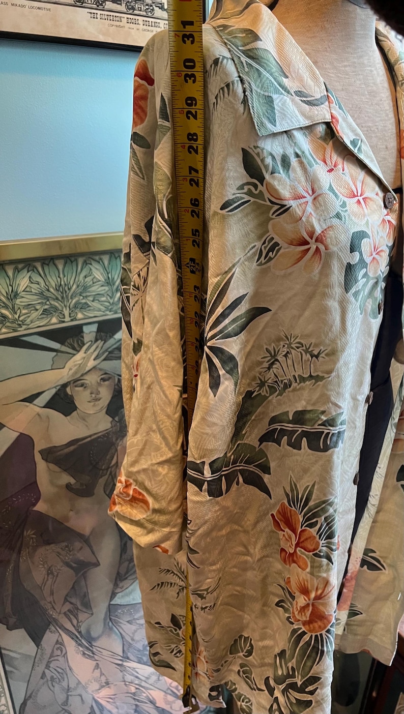 May include: A beige button-down shirt with a tropical floral print in orange, green, and white. The shirt is measured with a yellow tape measure, showing a length of approximately 25 inches.