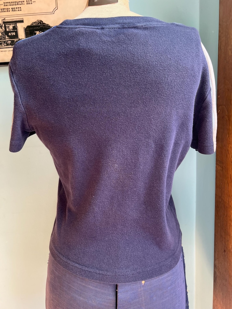 K&ouml;nnte beinhalten: Ein marineblaues Kurzarmshirt mit wei&szlig;en Streifen auf den &Auml;rmeln. Das Shirt wird auf einer Schaufensterpuppe pr&auml;sentiert. Der Stoff scheint ein weiches Strickmaterial zu sein. Das Shirt hat einen Rundhalsausschnitt.