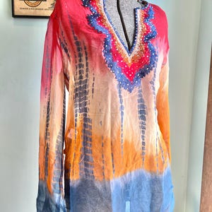 Peut inclure: Une tunique tie-dye aux tons de rouge, orange et bleu. Le haut &agrave; manches longues pr&eacute;sente une encolure en V avec des ornements perl&eacute;s. Le tissu semble transparent et l&eacute;ger, avec un motif tie-dye vertical.