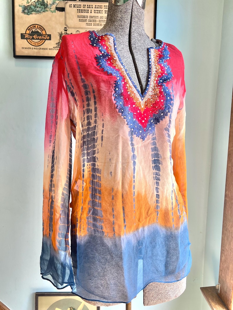 Peut inclure: Une tunique tie-dye aux tons de rouge, orange et bleu. Le haut &agrave; manches longues pr&eacute;sente une encolure en V avec des ornements perl&eacute;s. Le tissu semble transparent et l&eacute;ger, avec un motif tie-dye vertical.