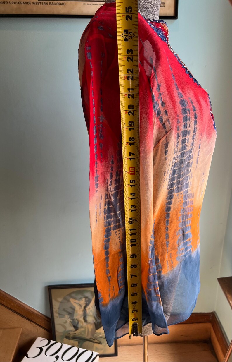 Peut inclure: V&ecirc;tement color&eacute; de style tie-dye, rouge, orange et bleu. Le tissu est drap&eacute; sur un mannequin, avec un m&egrave;tre ruban indiquant une longueur d'environ 63,5 cm. Le v&ecirc;tement a un aspect fluide et l&eacute;ger.