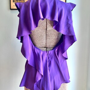 May include: A vibrant purple top with layered ruffles and a unique open back design. The garment is displayed on a mannequin, showcasing its flowing fabric and tailored silhouette. The top's color is a rich, saturated hue, suggesting a bold fashion statement.