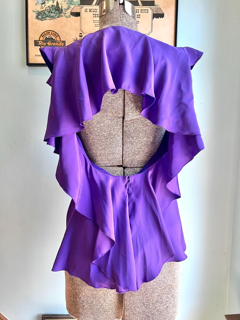 May include: A vibrant purple top with layered ruffles and a unique open back design. The garment is displayed on a mannequin, showcasing its flowing fabric and tailored silhouette. The top's color is a rich, saturated hue, suggesting a bold fashion statement.